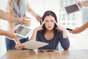 Stressed woman at a desk holding her head while multiple hands offer her devices and coffee.