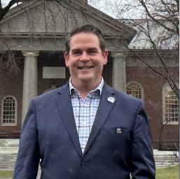 photo of professionally dressed man in suit standing in front of college building smiling
