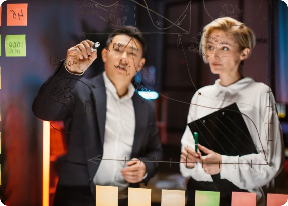 Two professionals writing diagrams and notes on a glass board during a strategy session