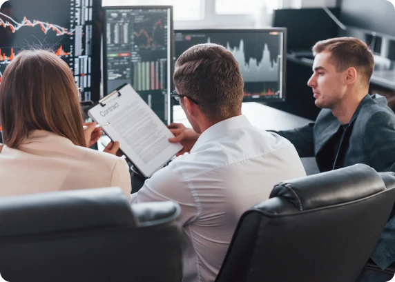 Three professionals reviewing contract documents and financial charts on computer monitors