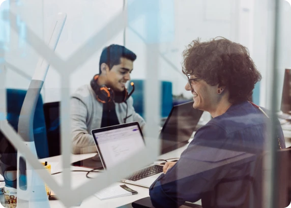 Two professionals smiling while working on laptops in a modern office behind glass
