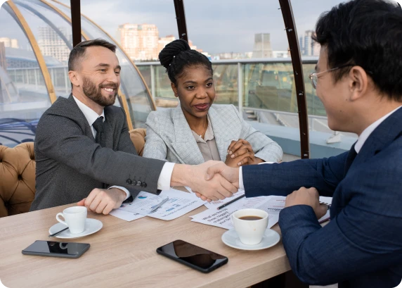 Three professionals shaking hands during a financial meeting at a table with documents and coffee