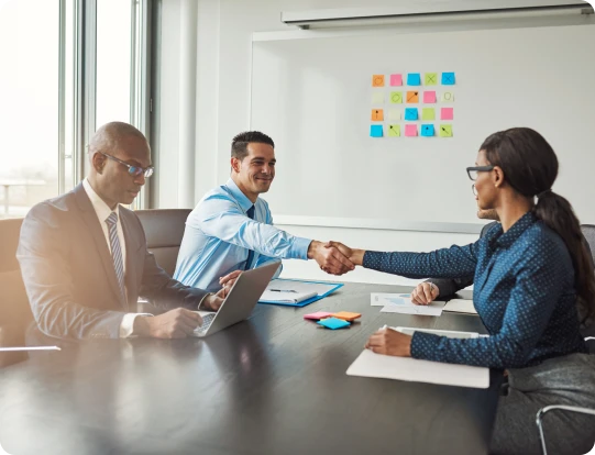 Three professionals in a conference room shaking hands across a meeting table